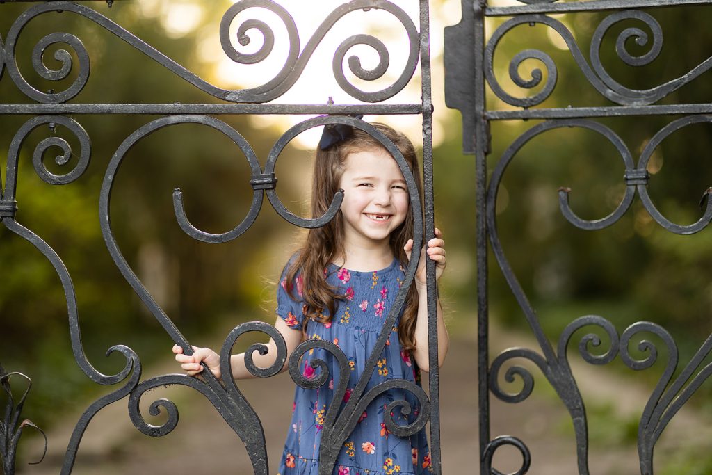 Five-year-old girl peeking through an iron gate, smiling proudly and showing off her missing teeth