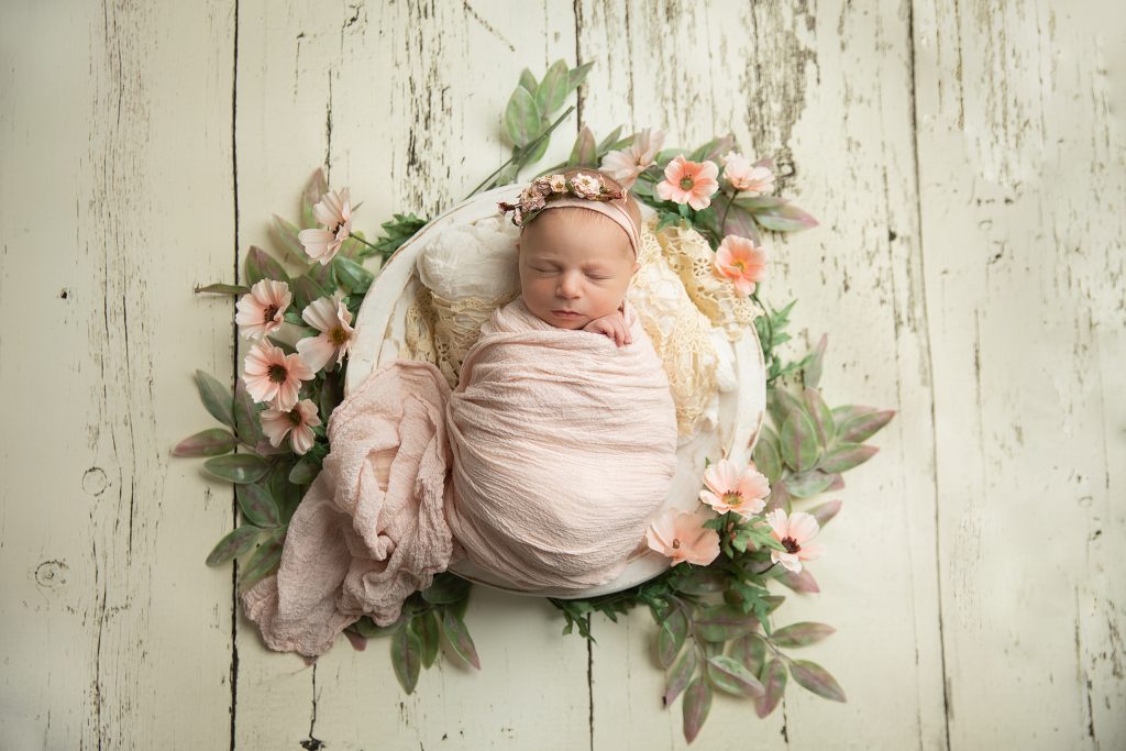 Newborn baby wrapped in pink with soft floral accents on a cream rustic wood floor, Delaware newborn photography session