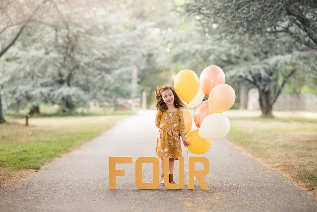 Four-year-old girl at Bellevue State Park in Wilmington, Delaware, celebrating her birthday with mustard yellow dress, balloons, and letters spelling "FOUR" during an outdoor photo session