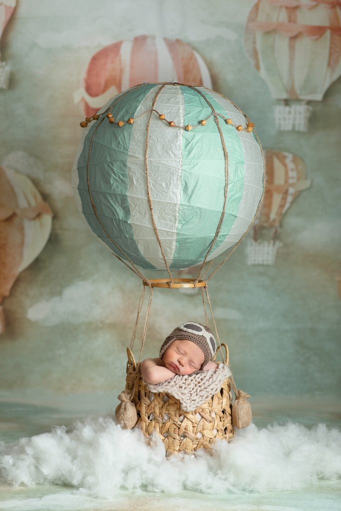 Newborn baby wearing aviator hat, posed in basket with hot air balloon theme props in soft mint green