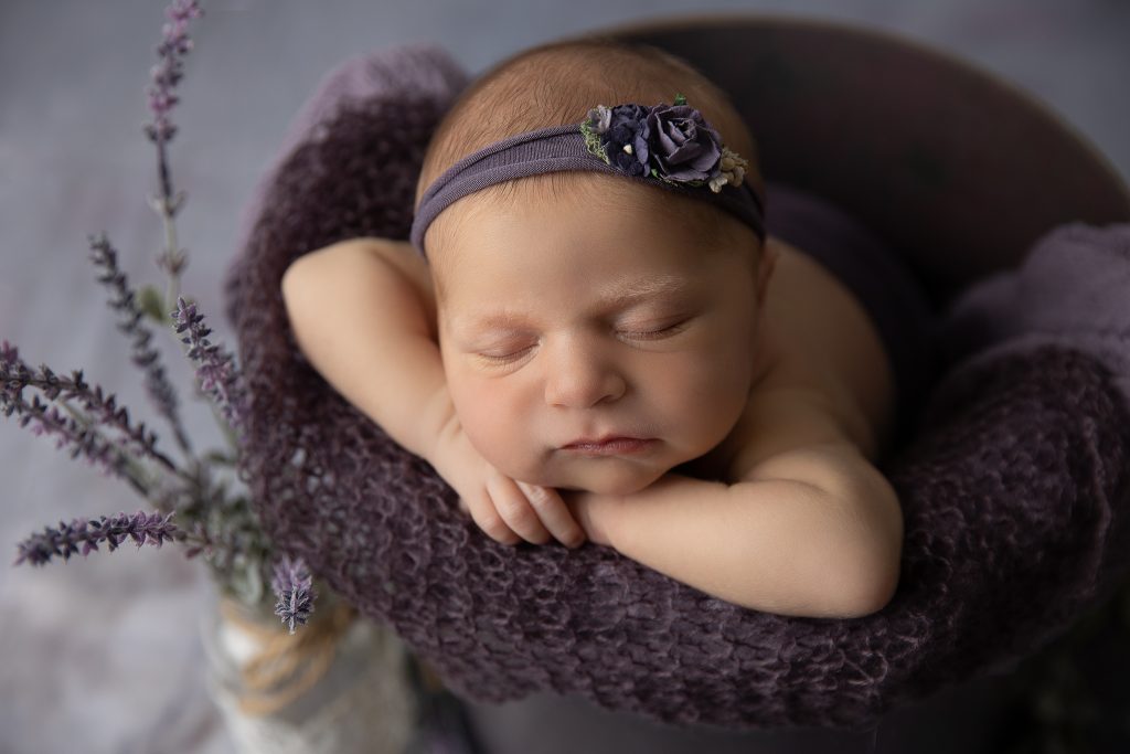 Newborn baby girl snuggled comfortably in a purple bucket with soft lavender flowers in a vase beside her