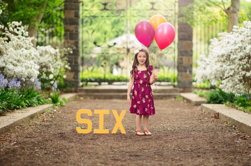 Six-year-old girl at her birthday photoshoot in Wilmington, Delaware, posing with pink and gold balloons and large “SIX” letters, with wrought iron gate in background