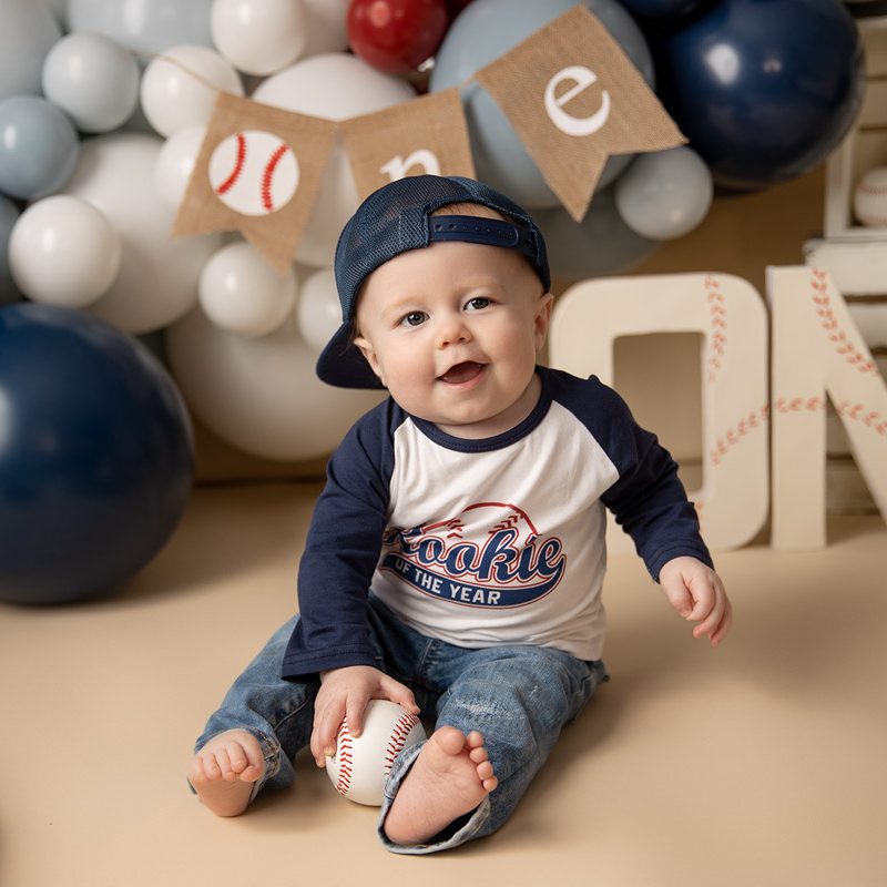 One-year-old boy in jeans and a baseball tee smiles while sitting among red, white, and blue balloons, vintage baseball props, and a “ONE” banner with baseball stitching for his Rookie of the Year first birthday session.