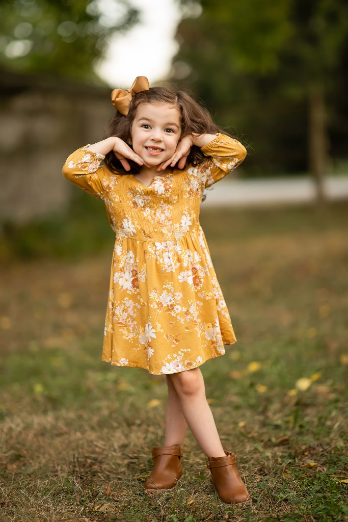Four-year-old birthday girl striking a sweet pose in a mustard yellow floral dress during an outdoor photo session.