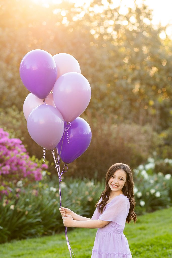 Nine-year-old girl in a lavender dress holding purple balloons, smiling in golden hour light during her outdoor birthday photoshoot