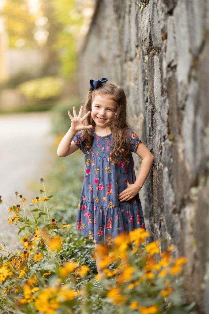 birthday girl standing near a stone wall surrounded by black-eyed susan flowers, holding up five fingers to show her age