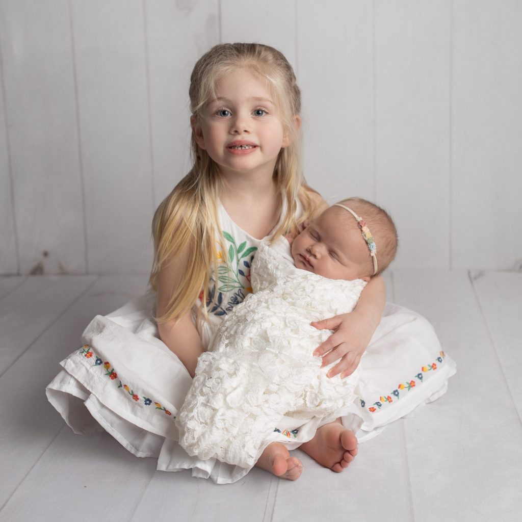 big sister holds newborn baby sibling in white lace wrap and pastel rainbow headband