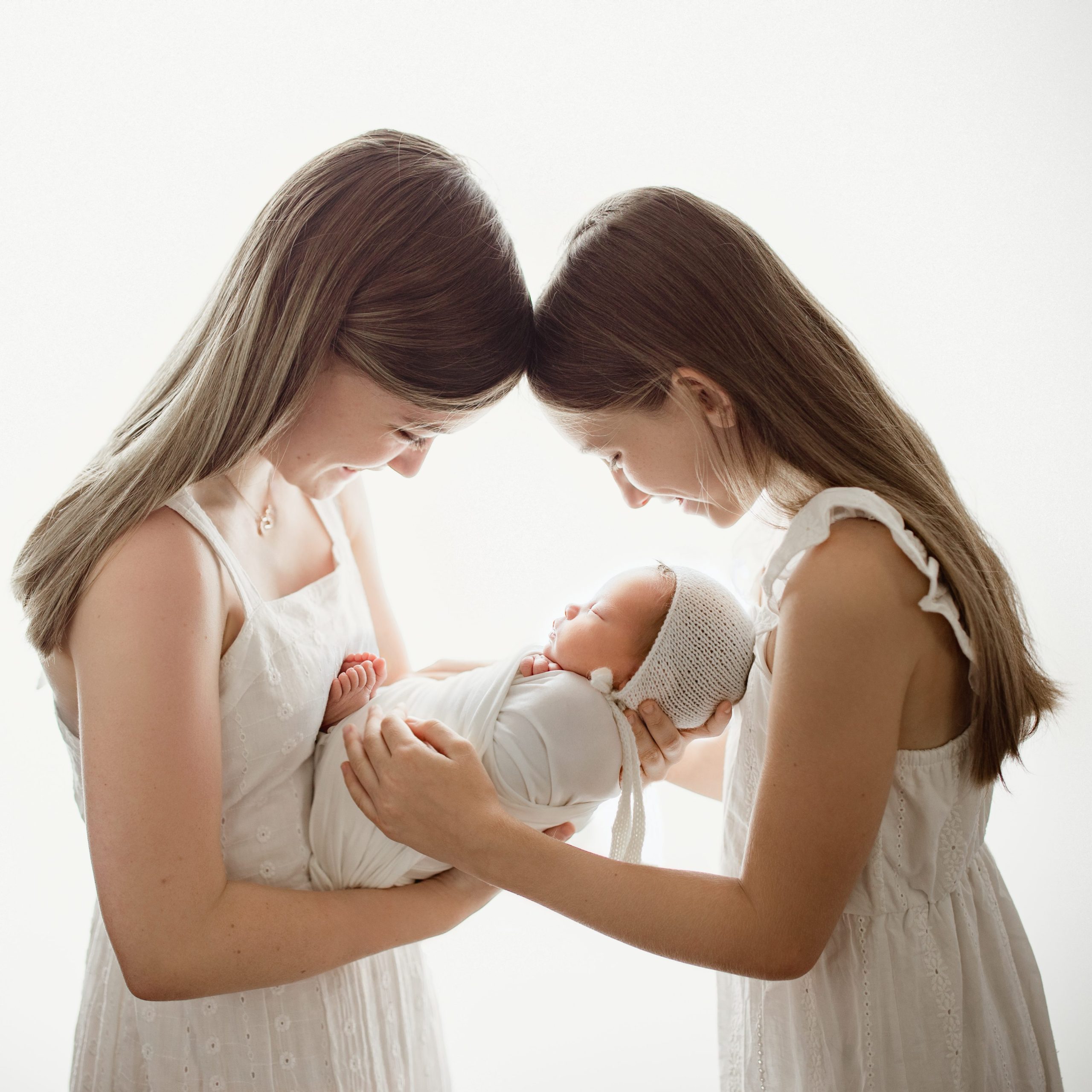 big sisters in white dresses holding baby sibling wrapped in a bonnet