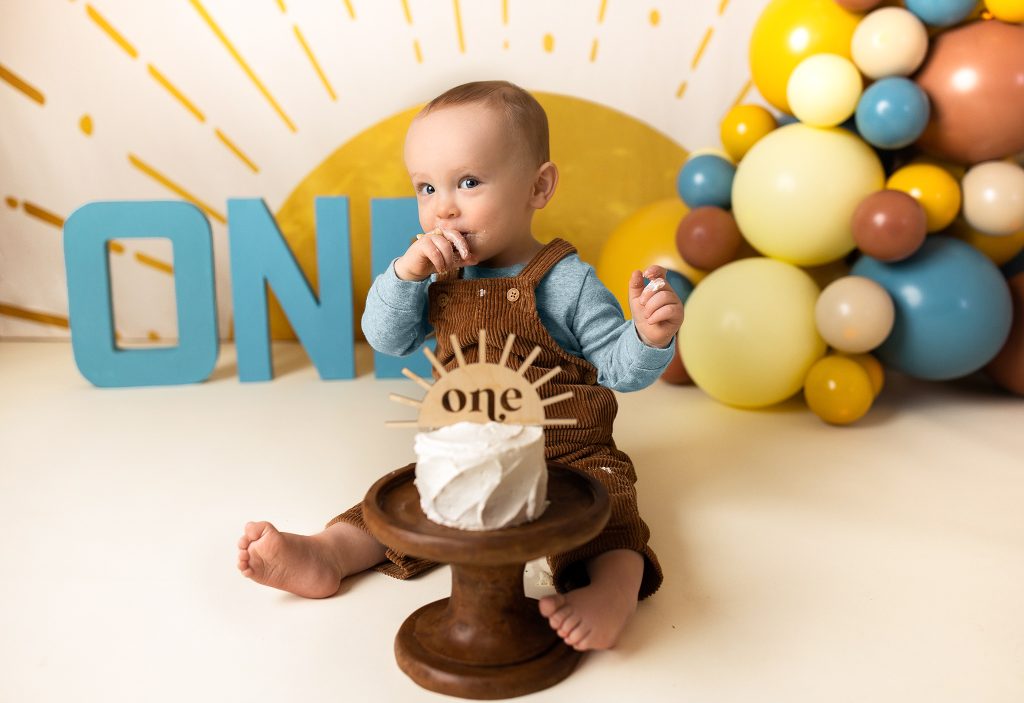 Baby gleefully digging into cake while seated, surrounded by warm-toned balloon garland and vintage sun accents.