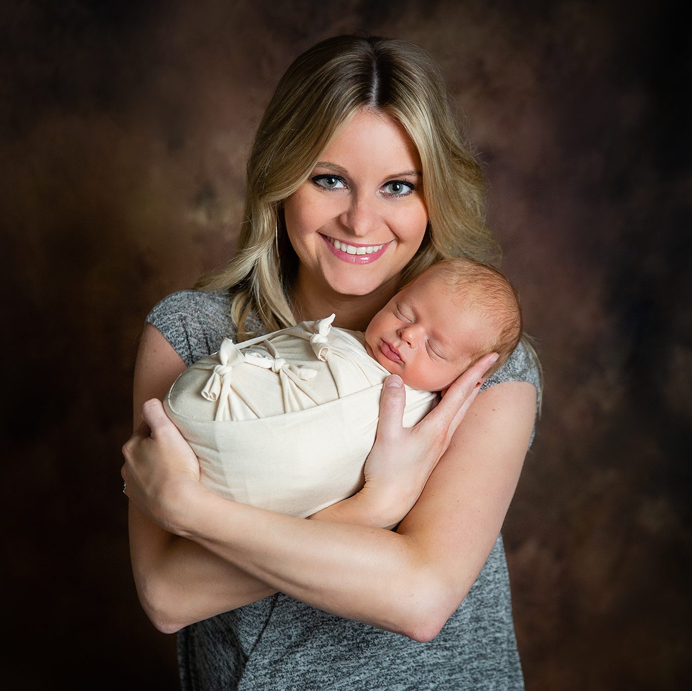 Portrait of a new mom holding her little boy wrapped in a cream swaddle.