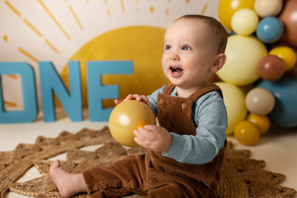 One-year-old sitting and holding a yellow balloon, smiling in front of a sunshine-themed backdrop.
