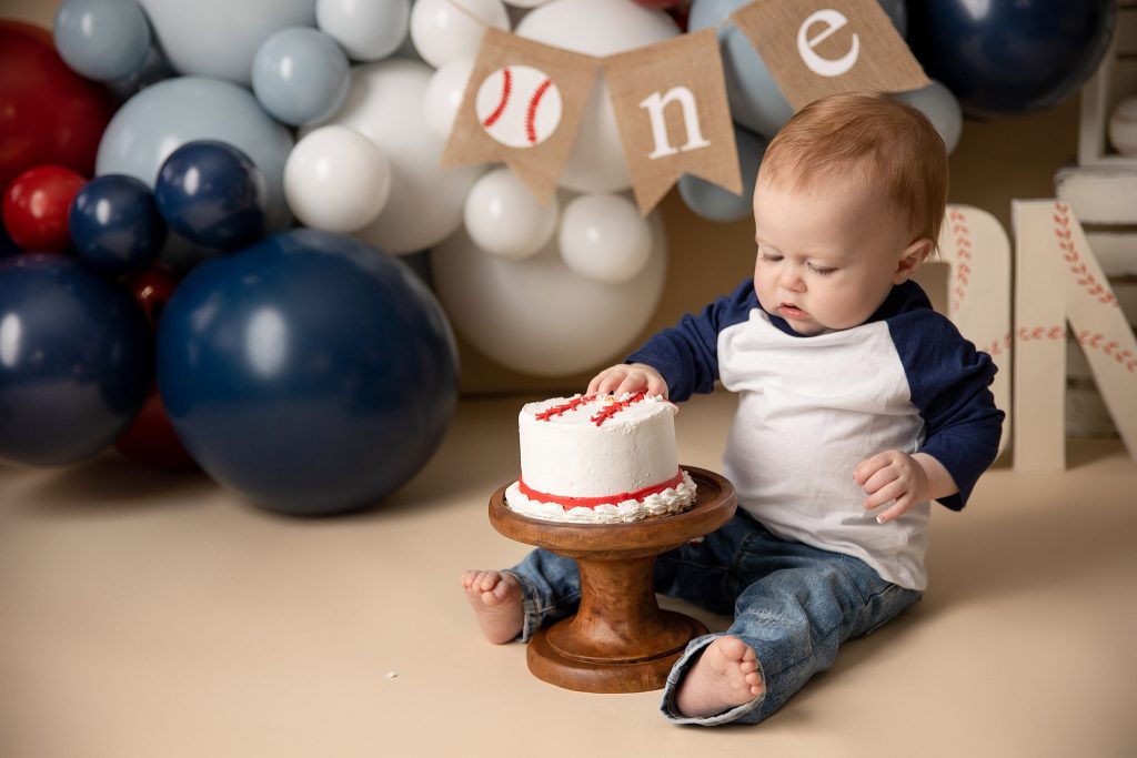 One-year-old boy in a vintage baseball birthday setup looks confused as he touches his cake for the first time, surrounded by themed props and balloons