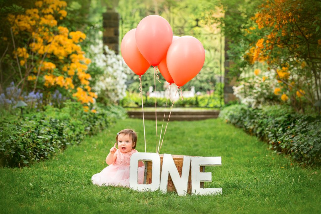 One-year-old celebrating their first birthday outdoors, holding up one finger with coral balloons and orange flowers in the background.