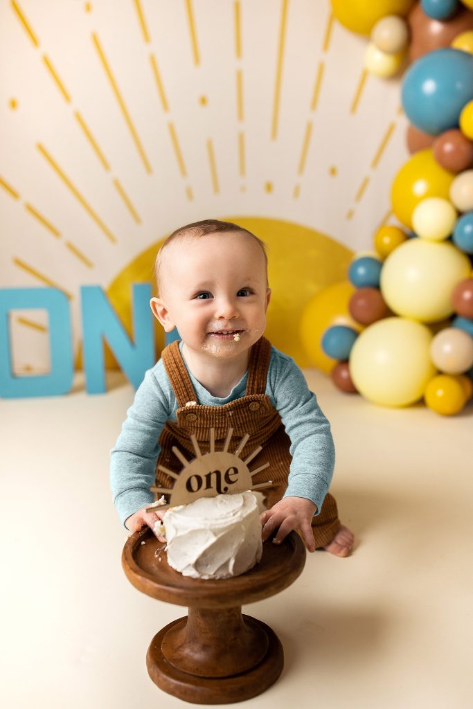 Toddler in overalls pushing a cake stand, cake smeared on hands and face.