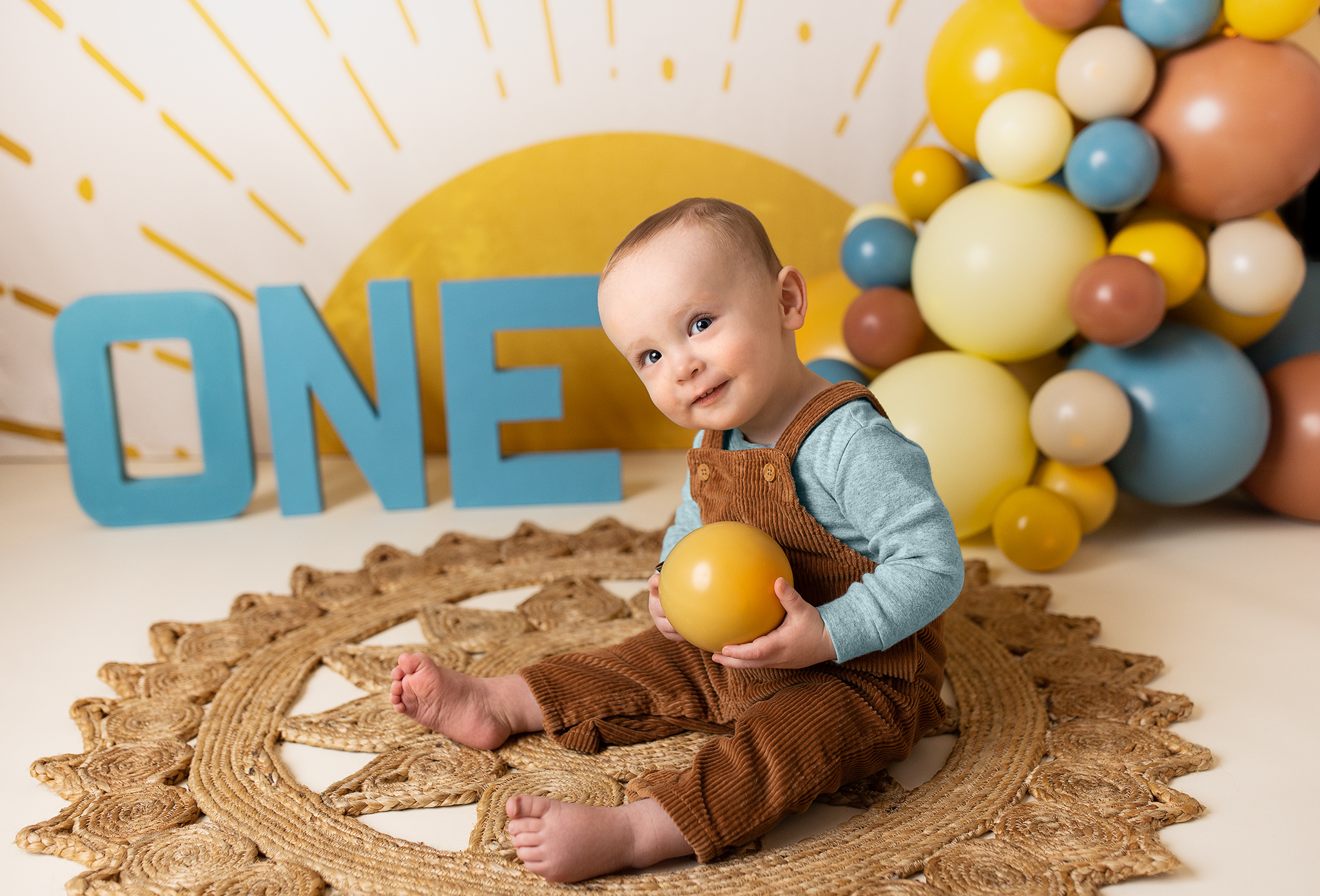 One-year-old sitting on jute rug and holding a yellow balloon, smiling in front of a 