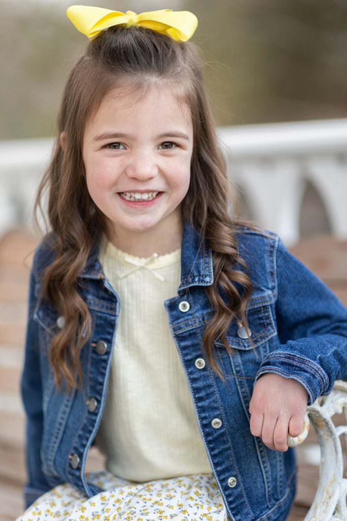 Five-year-old child wearing a yellow outfit and denim jacket, smiling brightly at the camera during an outdoor photo session