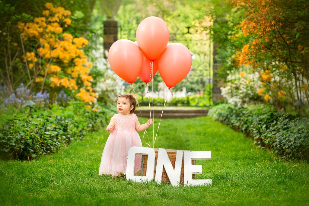 One-year-old standing next to coral balloons and large letters spelling ONE during a first birthday photo session