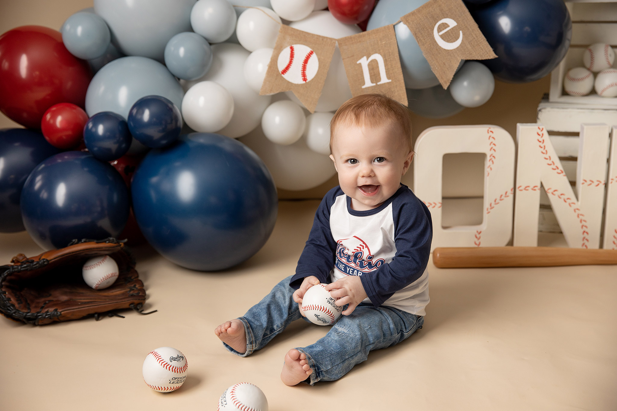 Smiling one-year-old in a baseball tee and jeans sitting with vintage baseball props and red, white, and blue balloon garland
