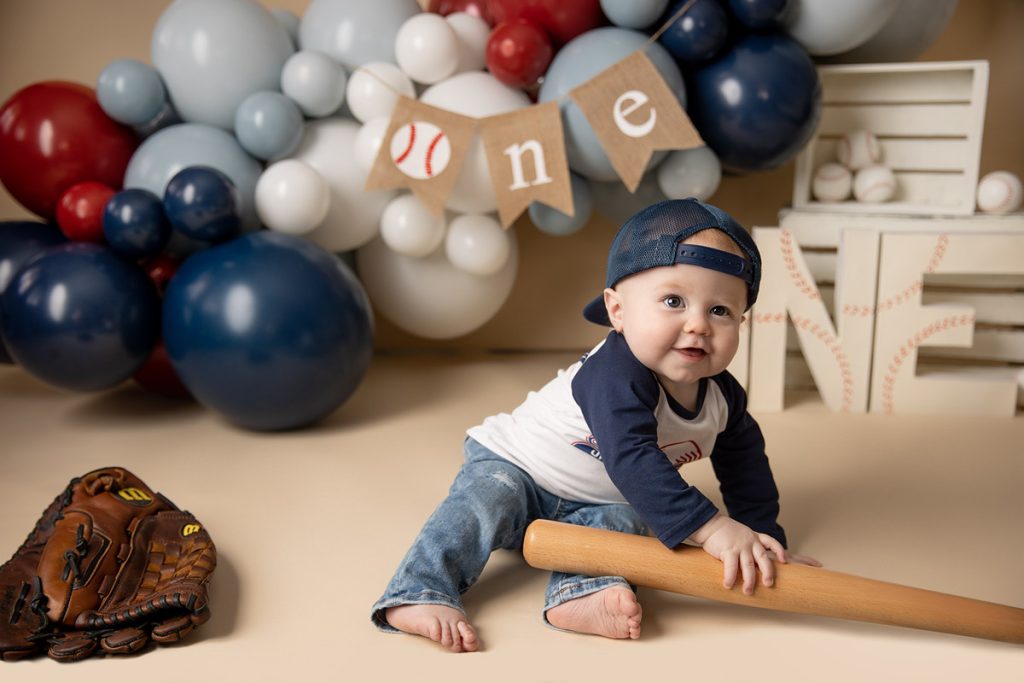 Smiling one-year-old in a vintage baseball-themed birthday setup with "Rookie of the Year" decorations, red, white, and blue balloons, and baseball props
