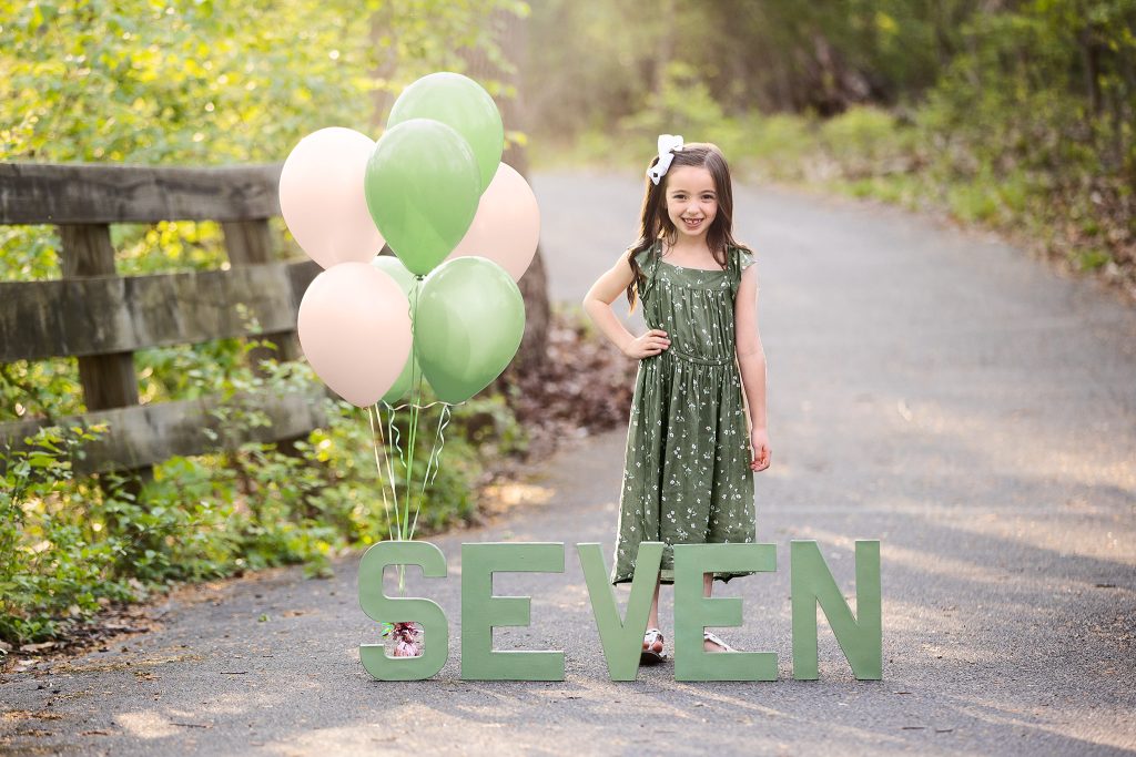 Seven-year-old girl celebrating her birthday outdoors, wearing a green dress with pink and green balloons and large letters spelling out "SEVEN" along a wooded path.