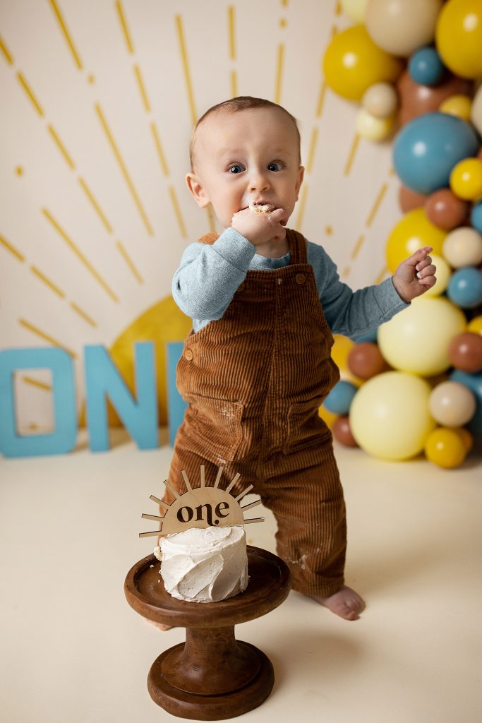 Baby standing behind a cake on wooden cake stand wearing brown corduroy overalls and a heather blue shirt, eating with frosting-covered fingers.