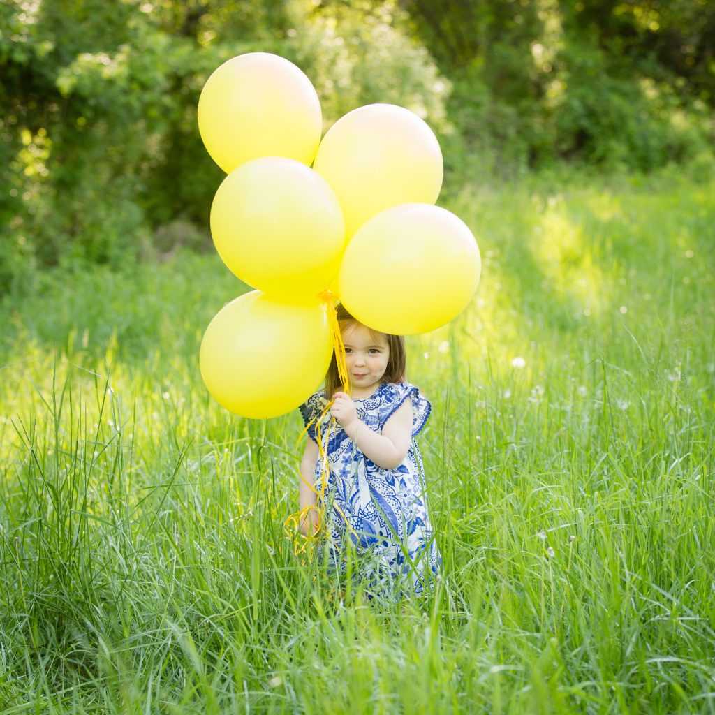 Two-year-old celebrating her birthday outdoors, playfully hiding behind bright yellow balloons in a sunlit field.
